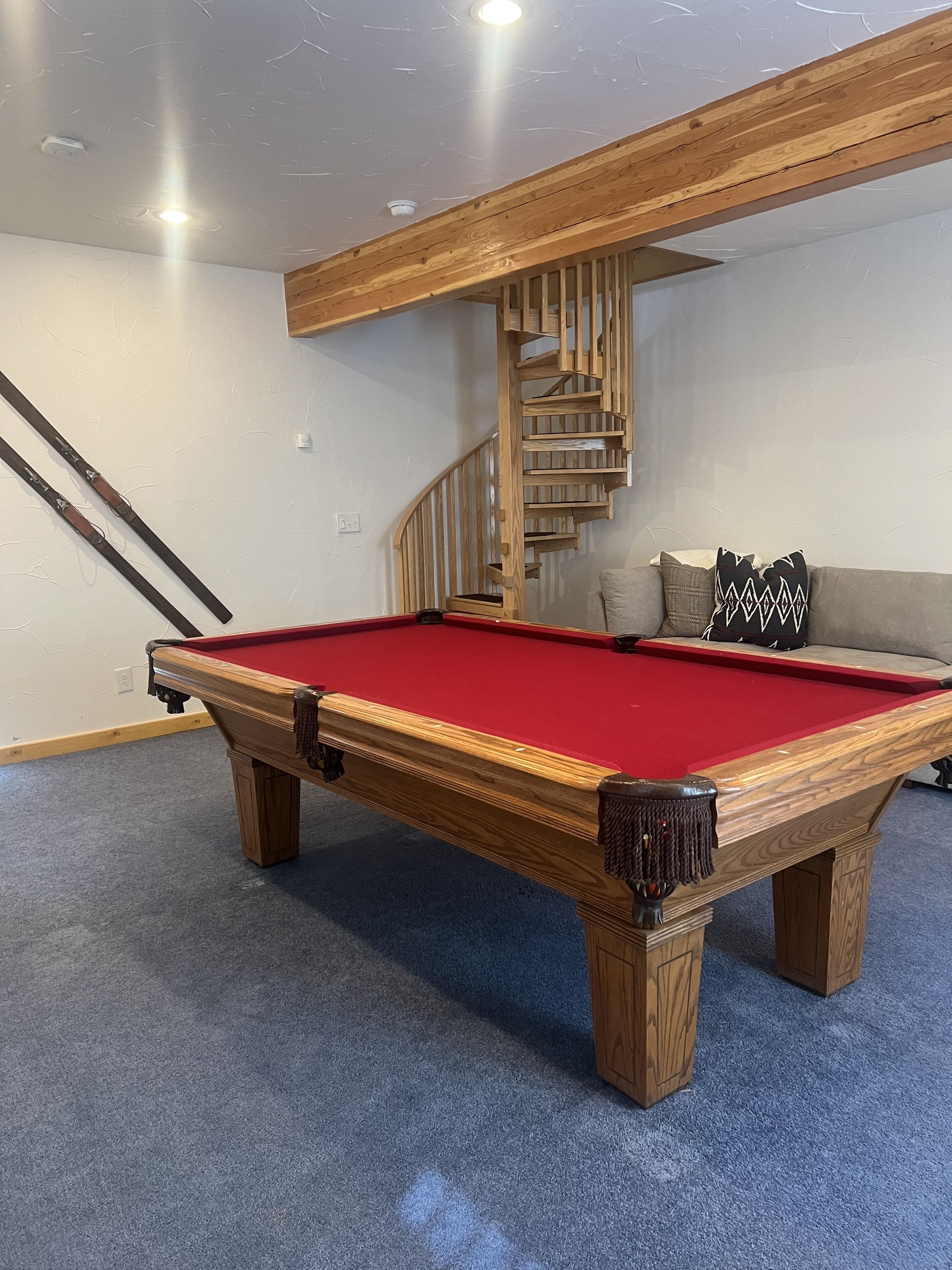 A pool table with a red felt surface, wooden legs, and a corner pocket. A wooden spiral staircase is visible in the background, leading to an upper level.