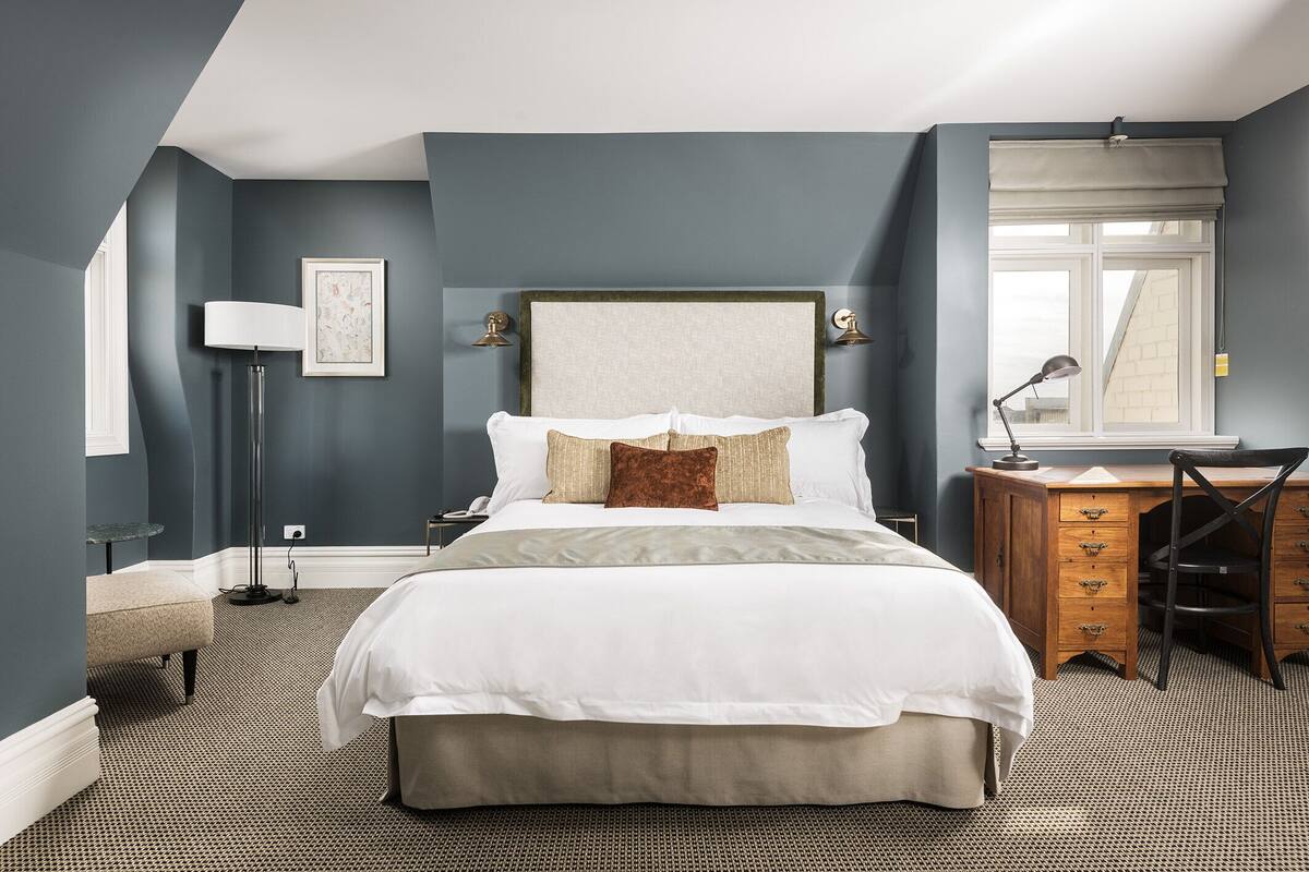 Une chambre à coucher avec un grand lit, un bureau, une chaise, une lampe et une fenêtre donnant sur un immeuble.