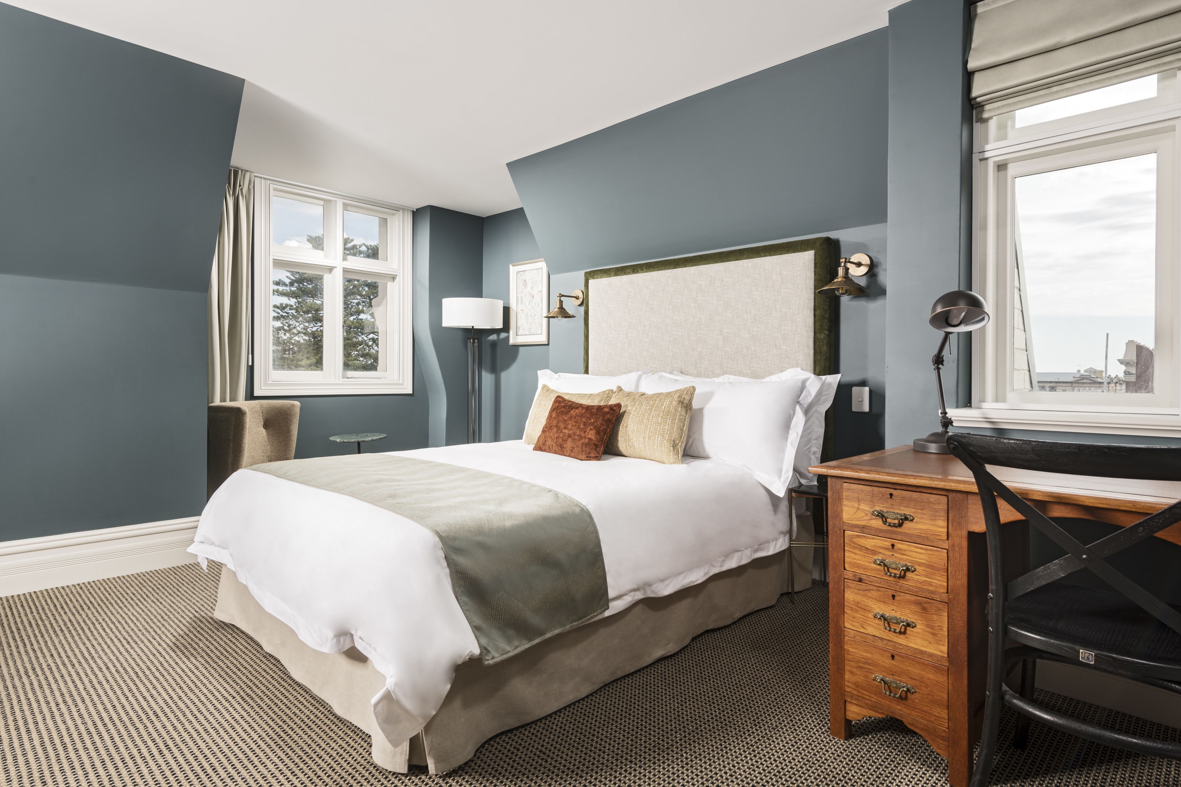 A neatly arranged bedroom with a bed, bedside table, lamp, and a desk with drawers.