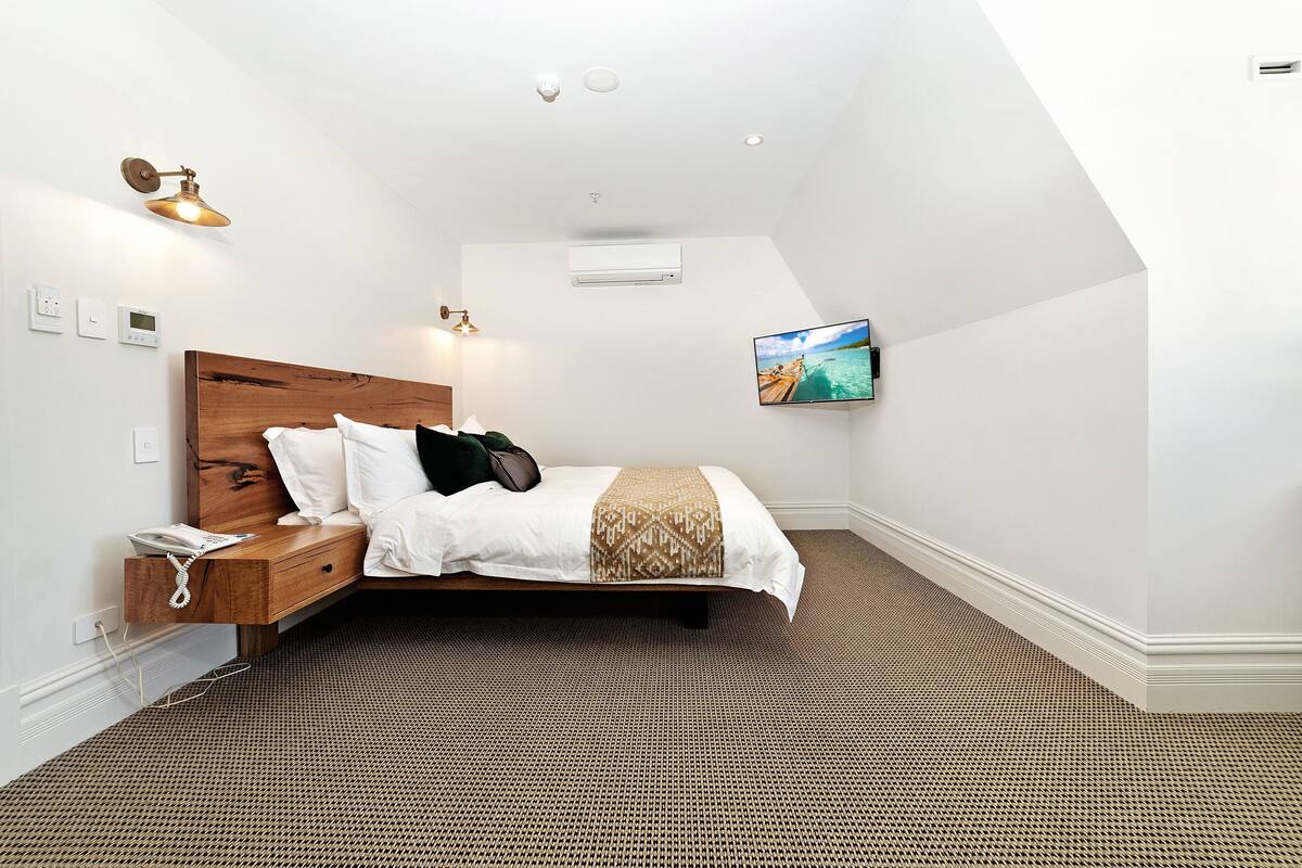 Une chambre moderne avec une tête de lit en bois, un lit recouvert d’une literie blanche et à motifs, une table de chevet avec un téléphone et un téléviseur fixé au mur.