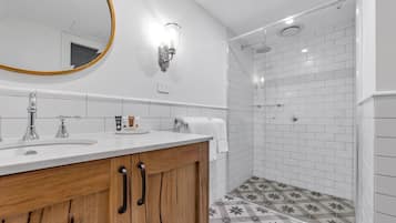 Un baño con tocador de madera, un espejo redondo, una lámpara de pared, ducha con azulejos blancos y piso estampado.