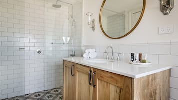 Un baño con paredes de azulejos blancos, un tocador de madera con lavabo, un espejo con marco dorado y una ducha con puerta de cristal.