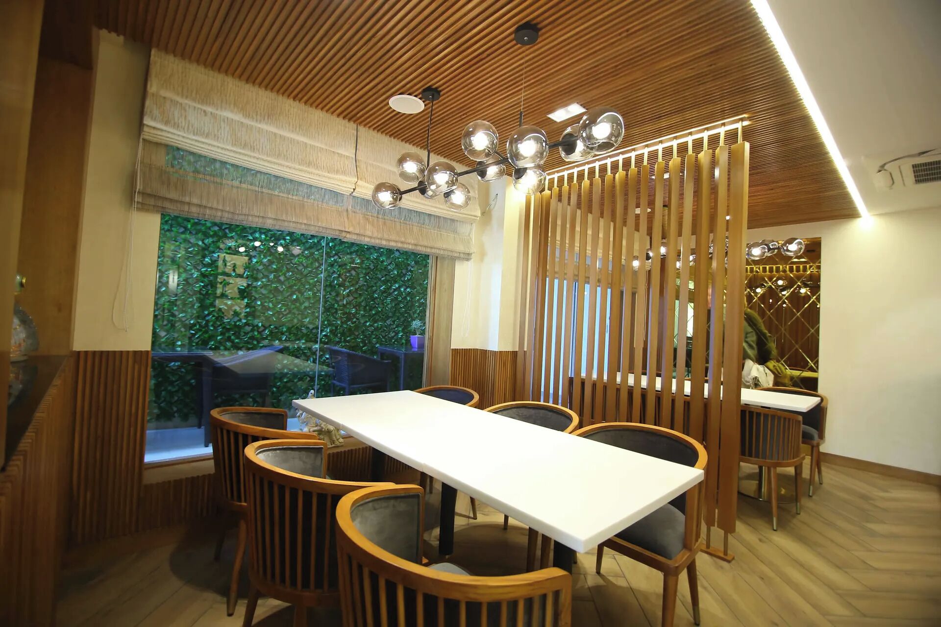 A restaurant interior with a long dining table, wooden chairs, a decorative ceiling, and a view through a glass wall.