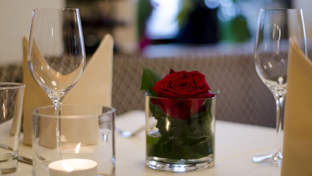 Une table à manger dressée avec de la vaisselle transparente, une bougie et un vase en verre contenant une rose rouge.