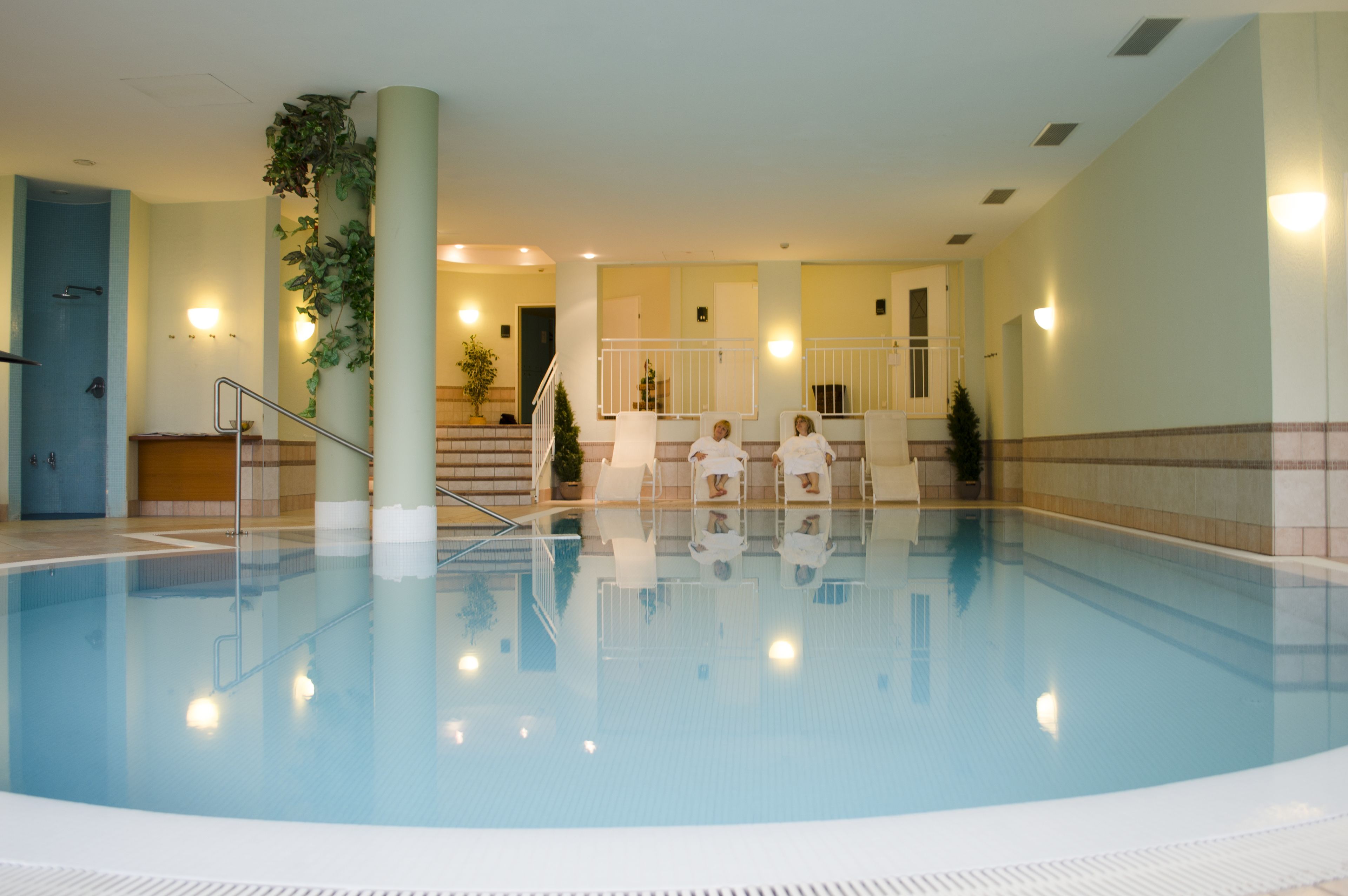 En indendørs swimmingpool med en klar vandoverflade, en central søjle og et siddeområde med stole og en lille plante.