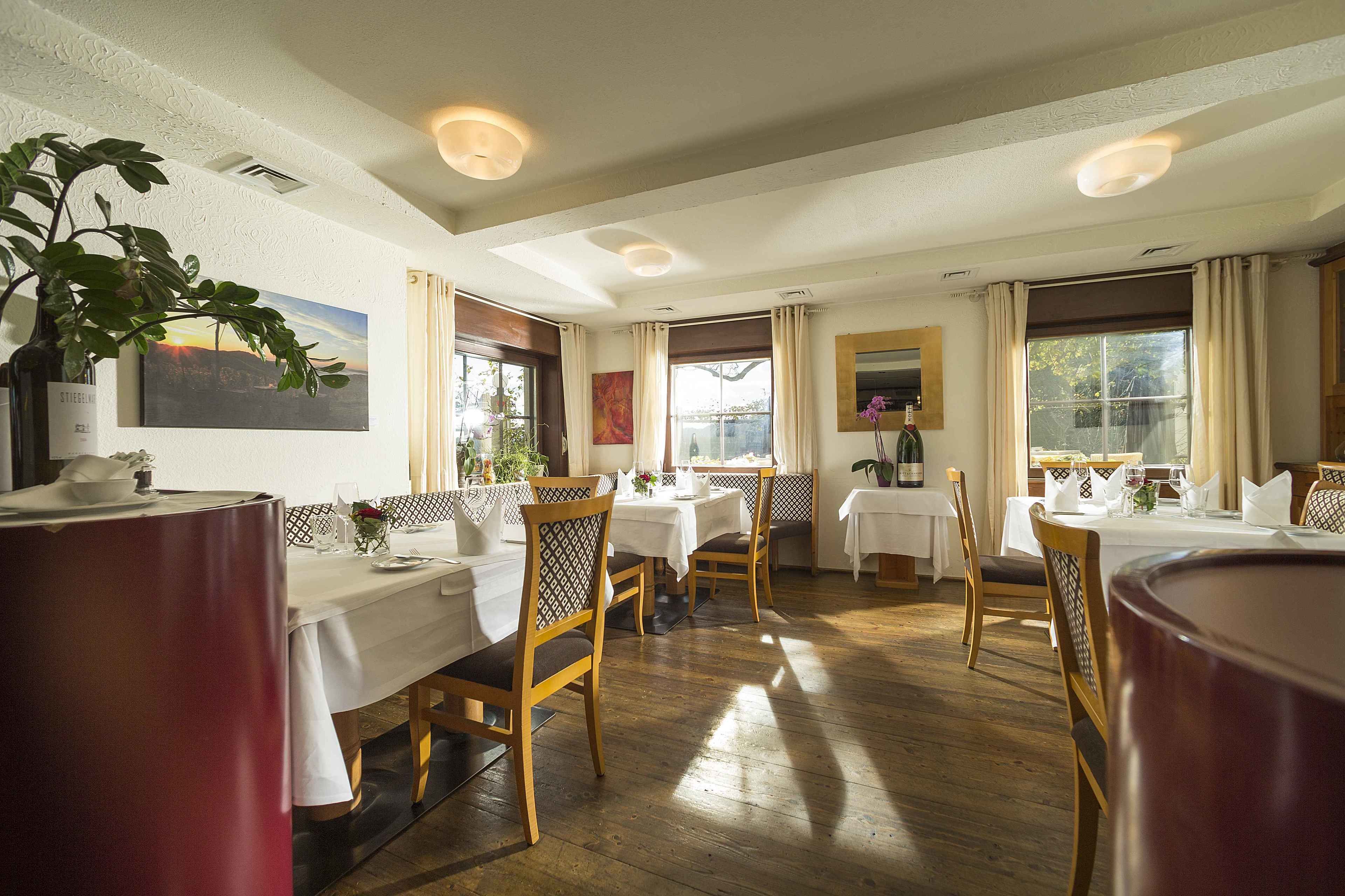 A dining area with wooden tables and chairs, white tablecloths, and a large potted plant.