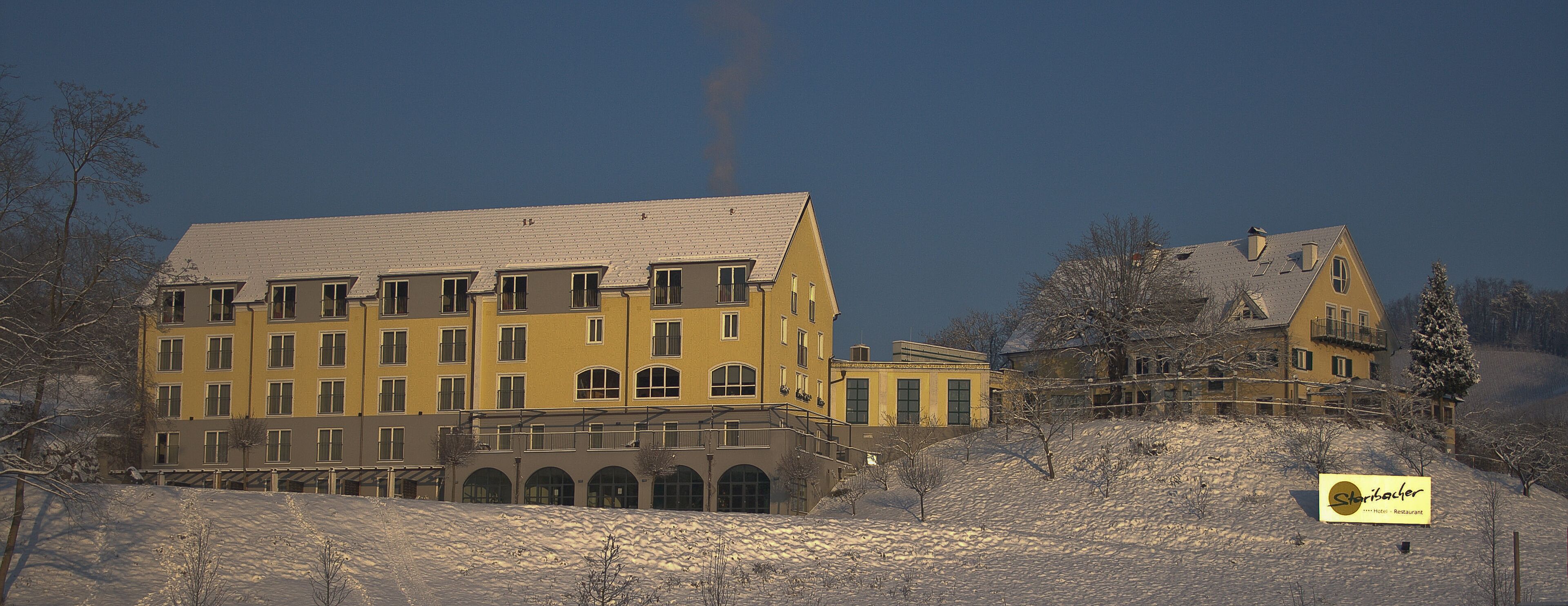 Un edificio amarillo grande con numerosas ventanas y un balcón, rodeado de árboles cubiertos de nieve y un cielo despejado.