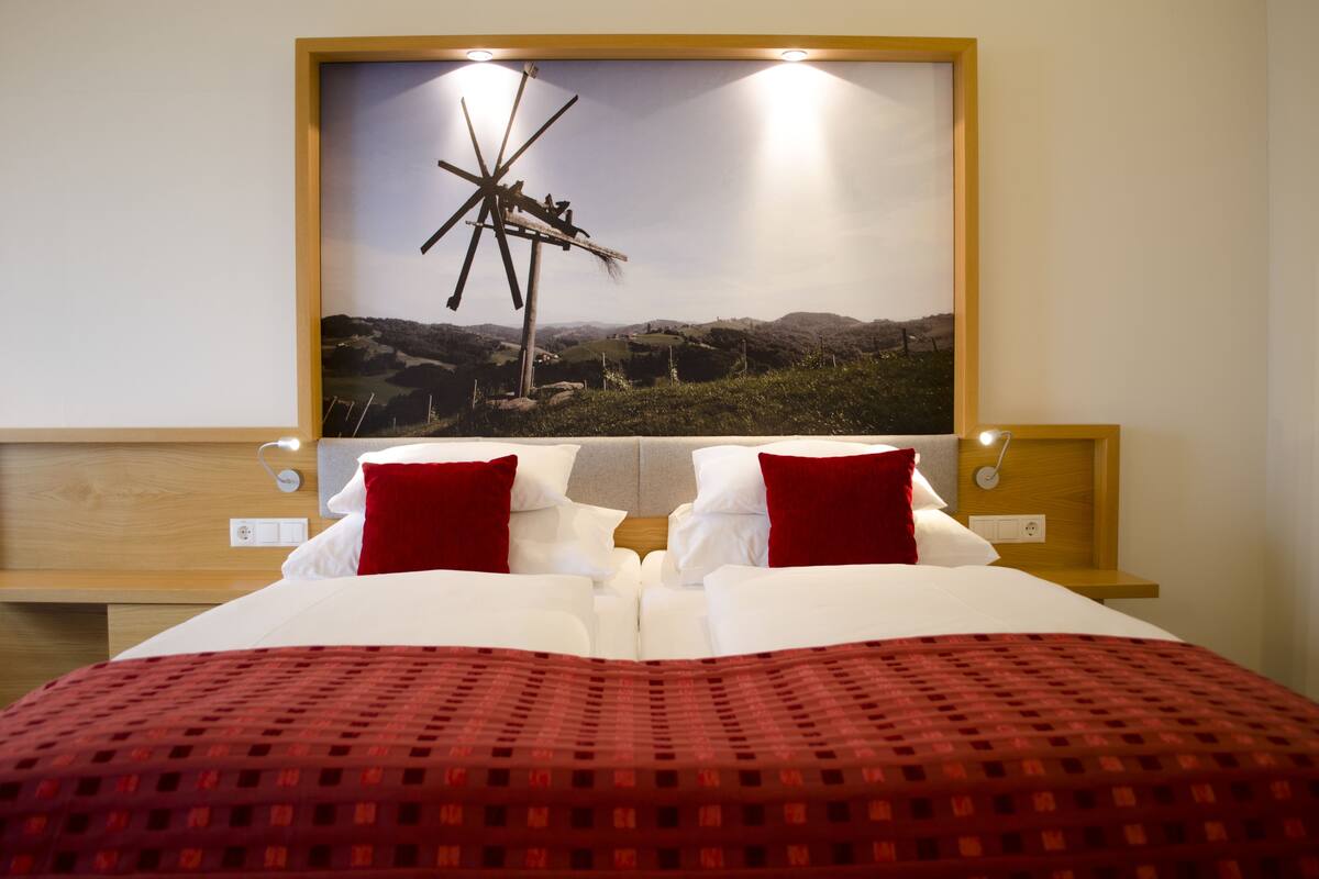A hotel room with a bed, two red pillows, a red patterned bedspread, a wooden headboard, and a large framed picture of a windmill on the wall.