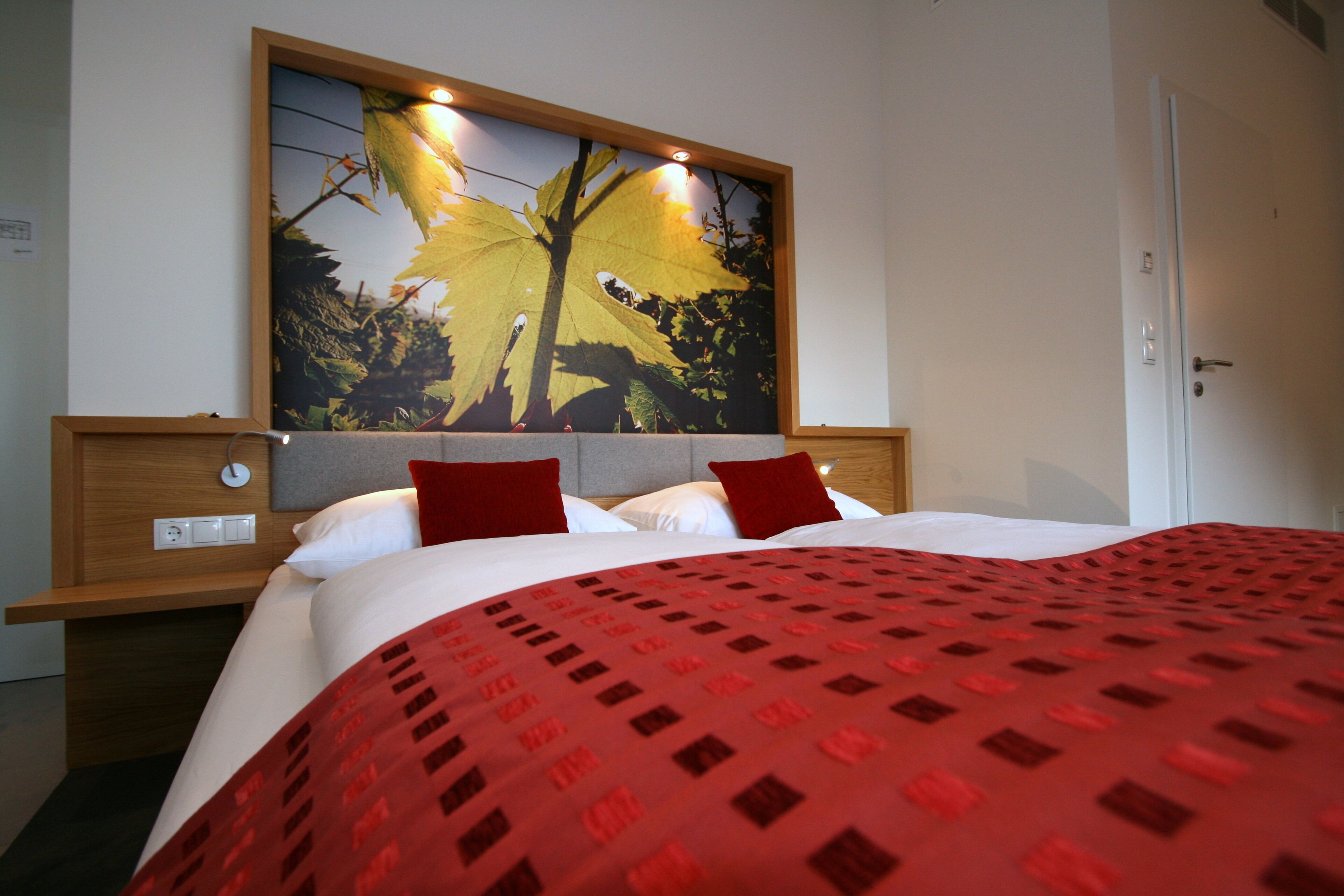 A hotel room with a large bed, two red pillows, a red patterned bedspread, a wooden headboard, a wall-mounted artwork of a tree, and a white door.