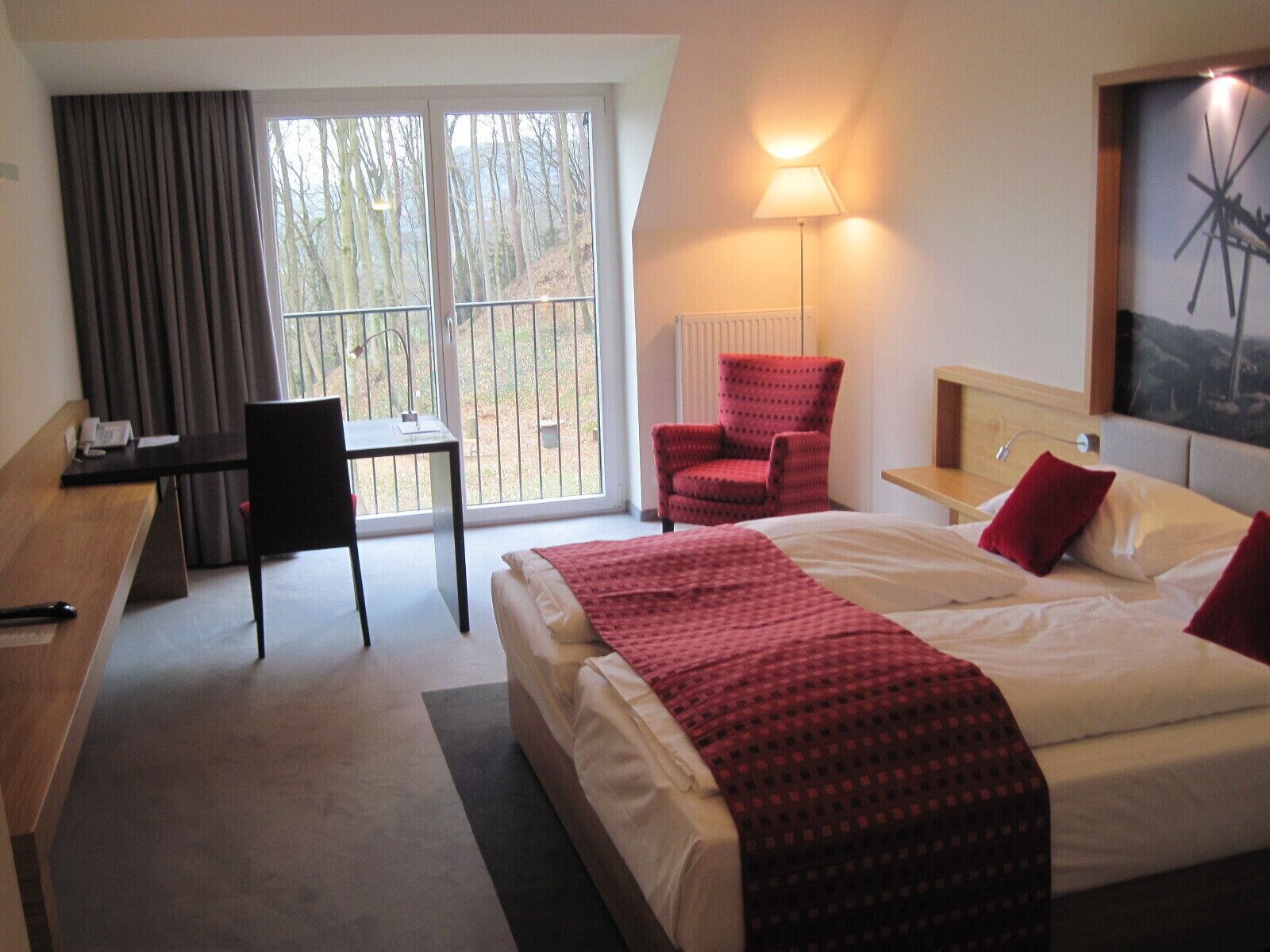 Habitación de hotel con cama, escritorio, silla, lámpara y ventana con vista a árboles.