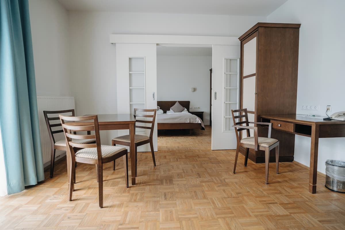 A room with a wooden herringbone floor, a bed with white bedding, a desk with a chair, and a wardrobe.