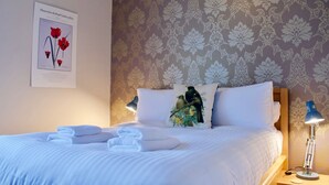 A neatly made bed with white linens and folded towels, a wall-mounted picture of flowers, and a patterned wallpaper. - Sophisticated Edinburgh Apartment in City Centre (Edinburgh)