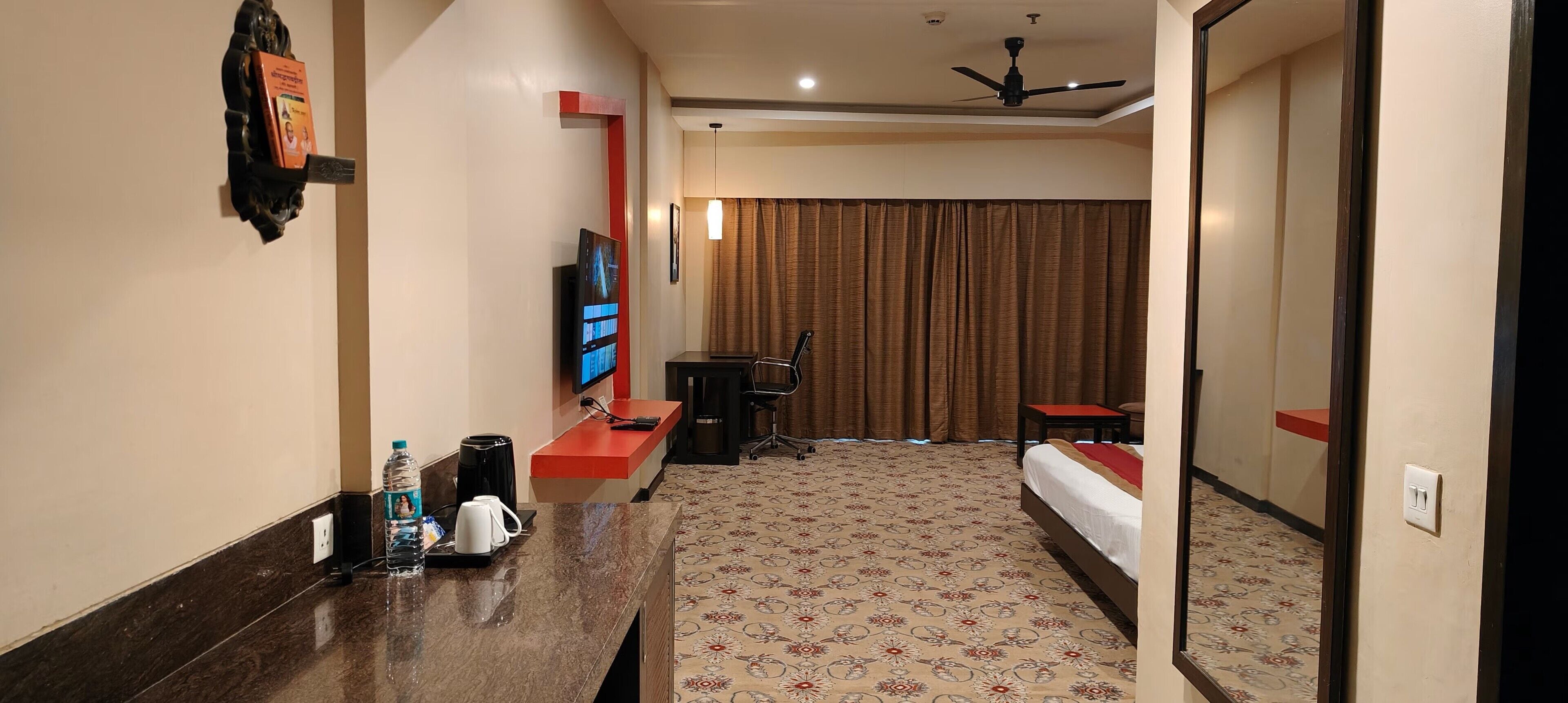 A hotel room with a bed, a desk with a TV, a chair, a small table with a kettle and cups, and a mirror.