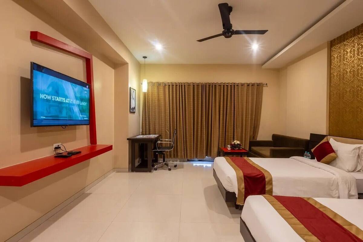 Ein Hotelzimmer mit Fernseher an der Wand, einem Schreibtisch mit Stuhl, zwei Betten mit roten und goldenen Bettdecken, einem Deckenventilator und einem Vorhang.