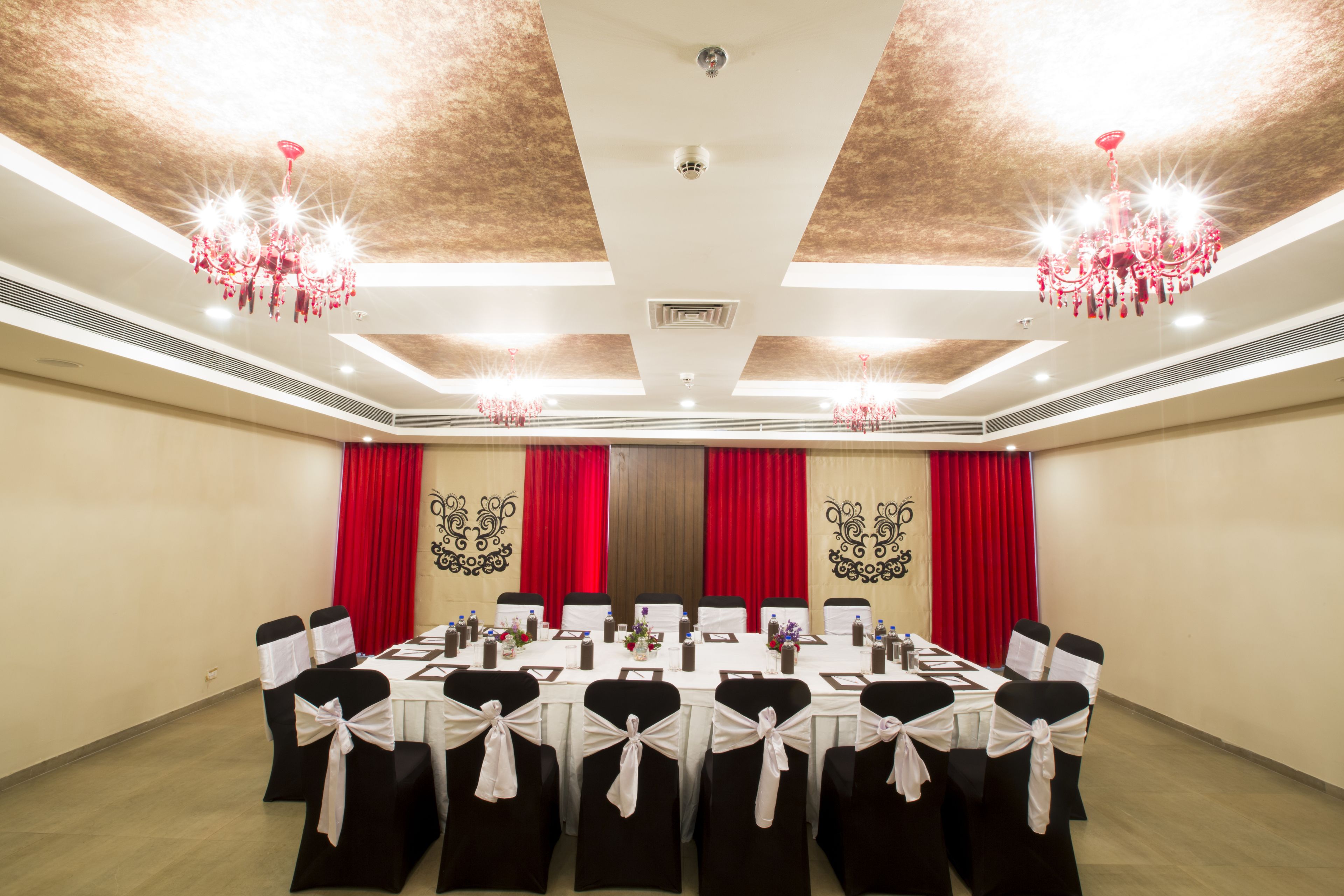 A conference room with a long table, black chairs with white sashes, red curtains, and chandeliers.