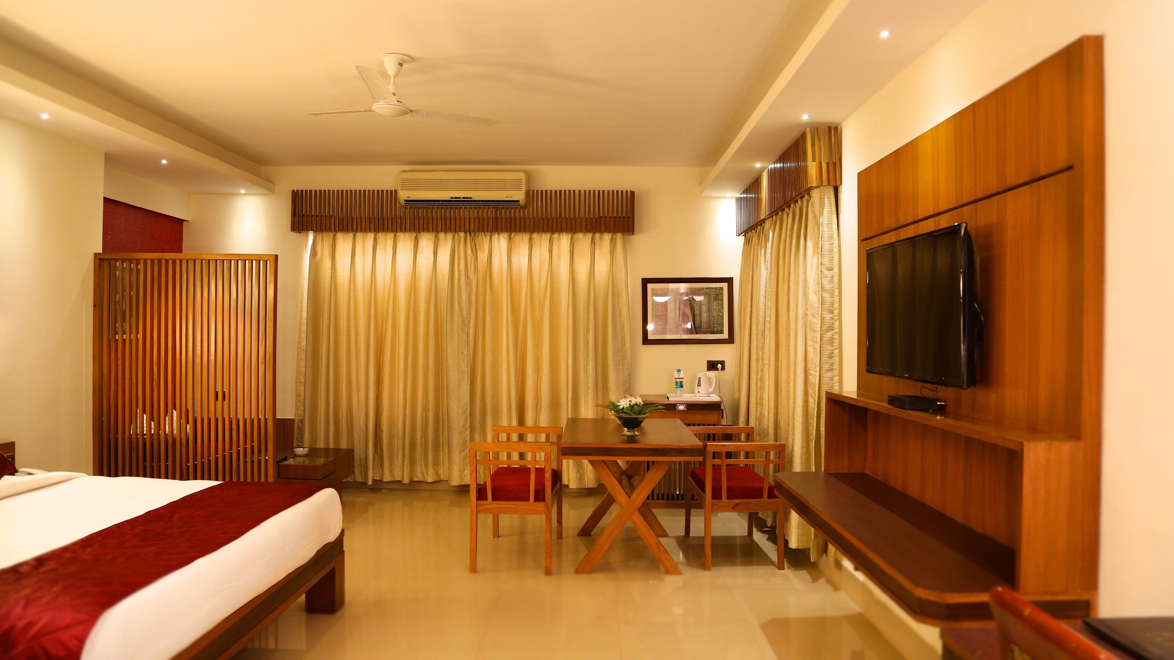 A hotel room with a bed, a desk with chairs, a television, an air conditioning unit, and a window with curtains.