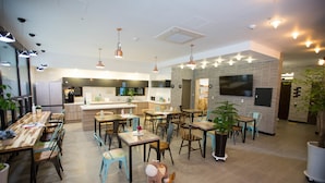 A modern dining area with wooden tables and chairs, a kitchen in the background, and a large TV screen on the wall. - Hotel Ggulzam (Yeosu)