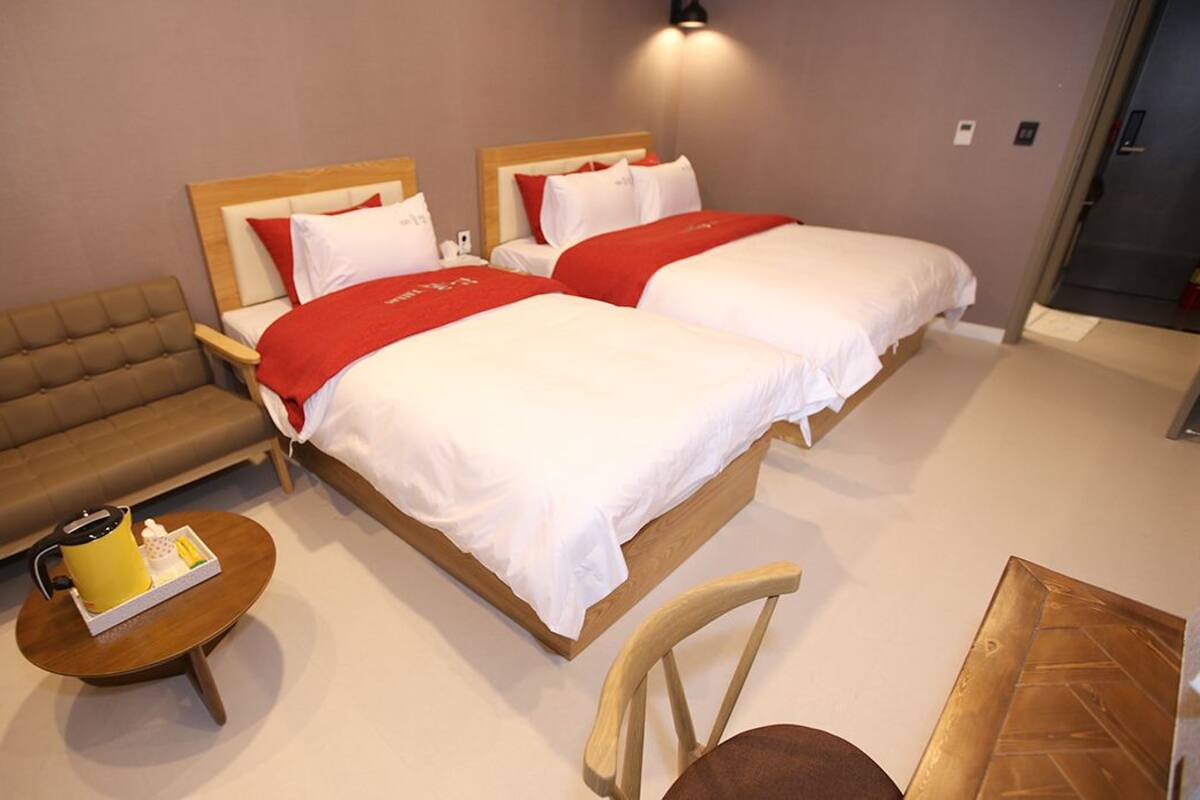 A hotel room with two beds, a sofa, a small table with a kettle, and a chair.