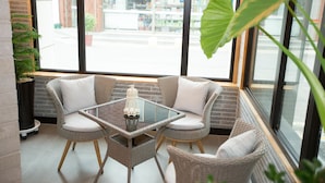 A corner seating area with wicker chairs and a glass table, a potted plant, and a view of a brick building outside. - Hotel Ggulzam (Yeosu)