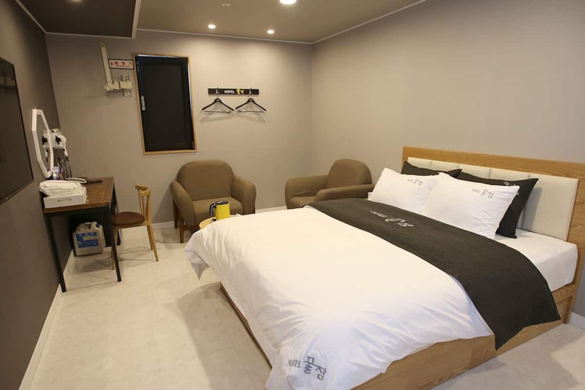 A hotel room with a large bed, two armchairs, a desk with a chair, a mirror, and a coat rack.