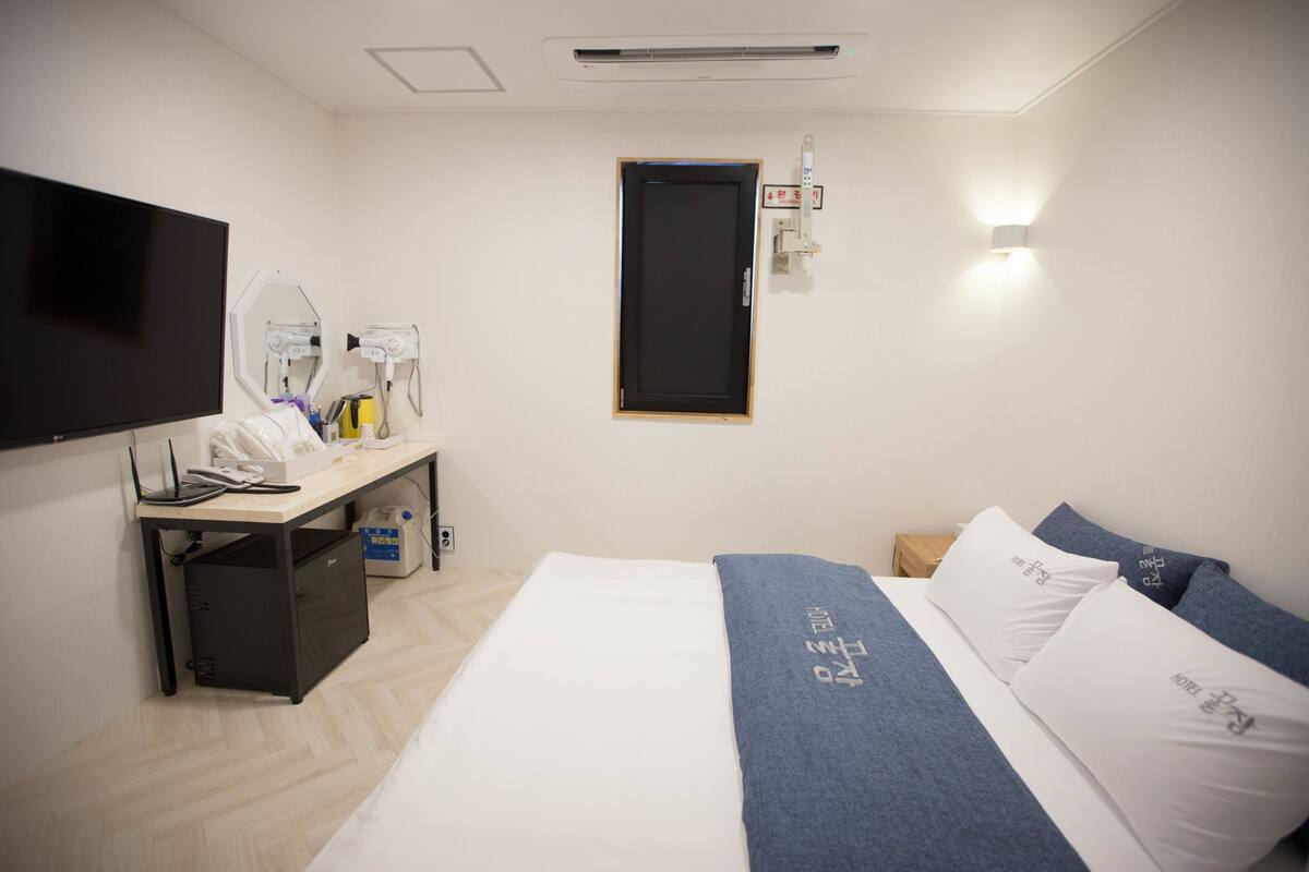 A hotel room with a bed, a desk with a computer, a TV, and a wall-mounted light.