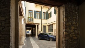 A narrow alleyway with a blue car parked on the right, stone walls, and a building with green shutters in the background. - Massimago Wine Suites (Verona)