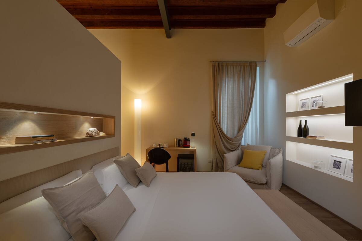A hotel room with a large bed, a desk with a chair, a TV, and shelves with books and decorative items.
