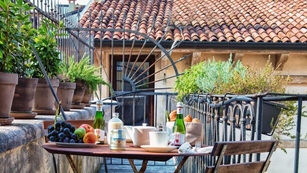An outdoor dining area with a table set for two, surrounded by a wrought iron fence, and a view of a building with a tiled roof. - Massimago Wine Suites (Verona)