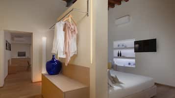 A modern bedroom with a bed, bedside tables, and a TV. There is a hallway leading to a kitchen area with a TV and a small seating area.