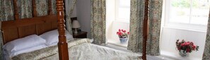 A four-poster bed with a floral bedspread, a bedside table with a lamp, and a window with a view of greenery. - OYO Rose & Crown Hotel (Leyburn)