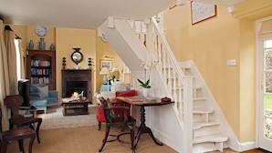 A cozy living room with a fireplace, a bookshelf, a dining table, and a staircase. - Saratoga Cottage (Chipping Norton)
