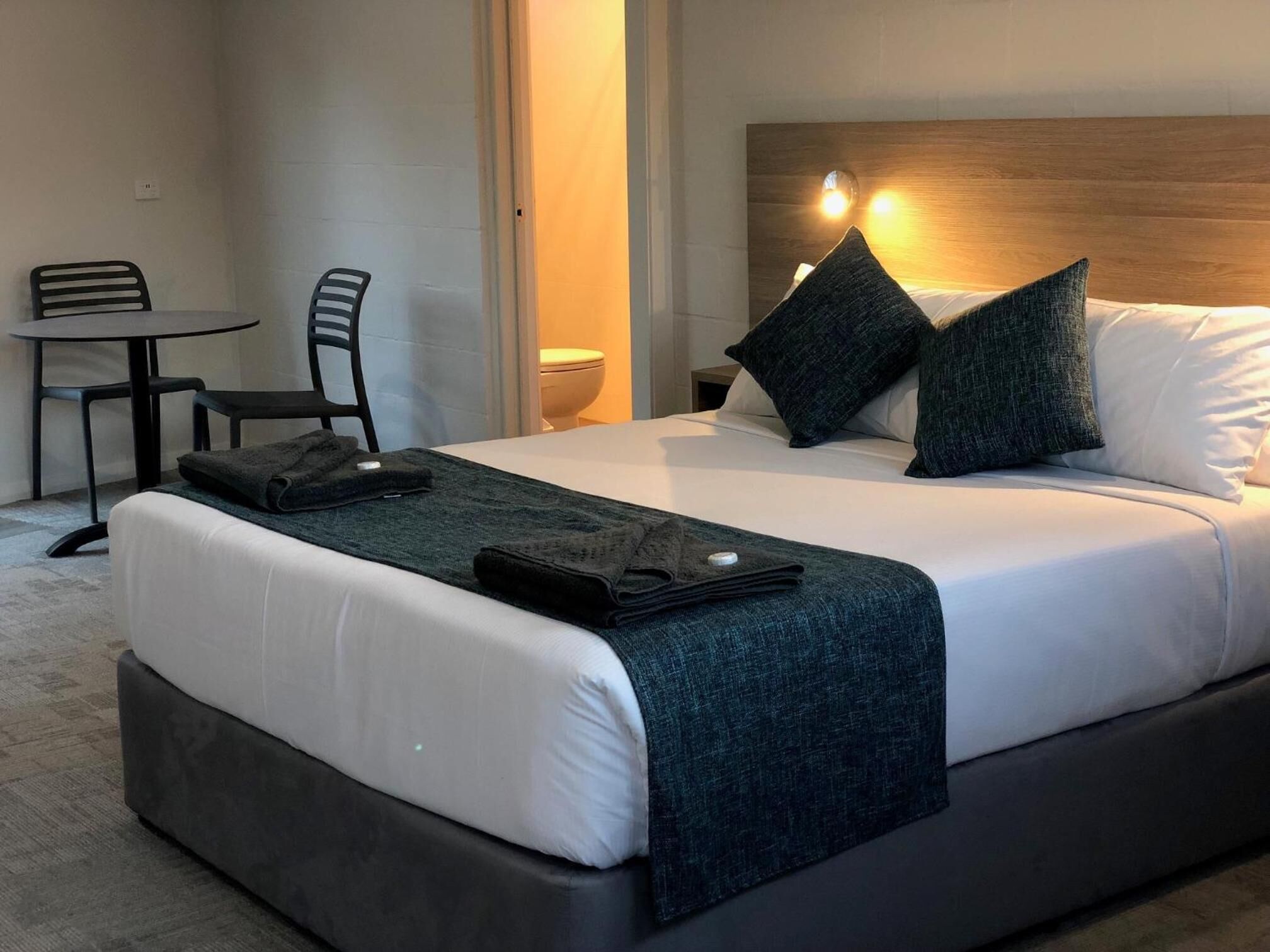 Habitación de hotel con una cama, una mesita, dos sillas, una mesita de noche y una lámpara de pared.