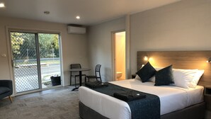 Quarto de hotel moderno com uma cama grande, uma mesinha e cadeiras, uma porta de vidro deslizante para uma área externa e uma cabeceira de madeira com luminárias fixadas na parede.