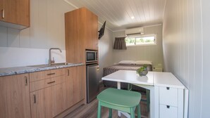 Cozinha compacta com armários de madeira, pia, micro-ondas e geladeira. Há uma pequena área de refeições com uma mesa branca e um banquinho verde. Uma cama com janela e cortinas é visível ao fundo.