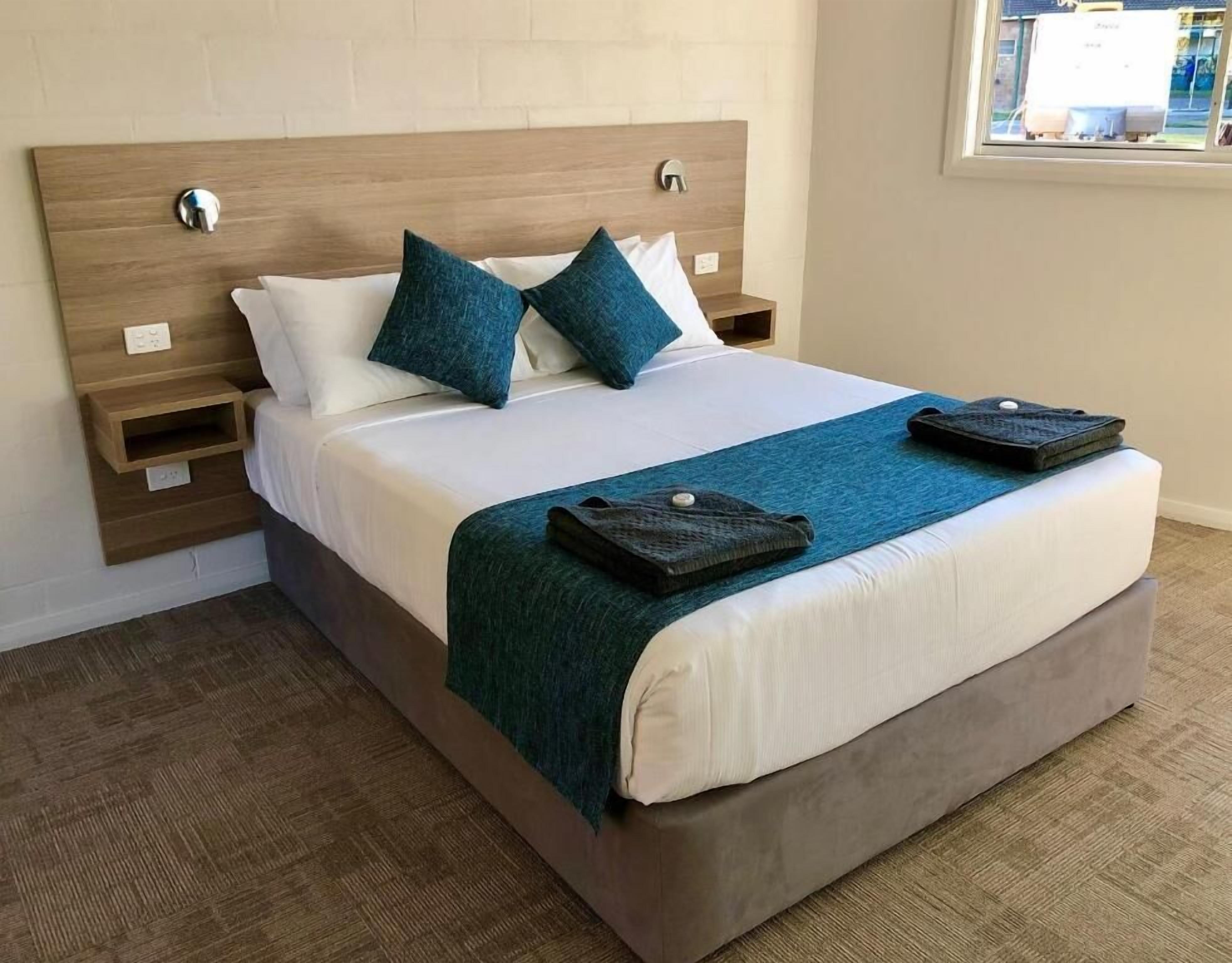 Un lit bien fait, avec une tête de lit en bois, deux oreillers, un plaid bleu et une petite table de chevet sur laquelle se trouve une lampe.