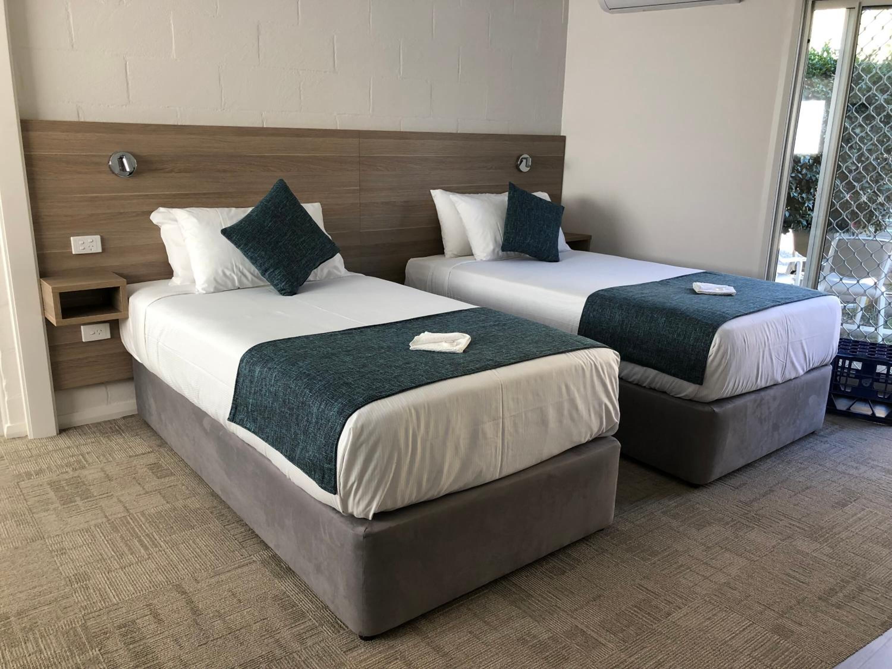A hotel room with two beds, each with a wooden headboard, white bedding, and teal throw pillows. There is a small wooden nightstand with a lamp and a power outlet.