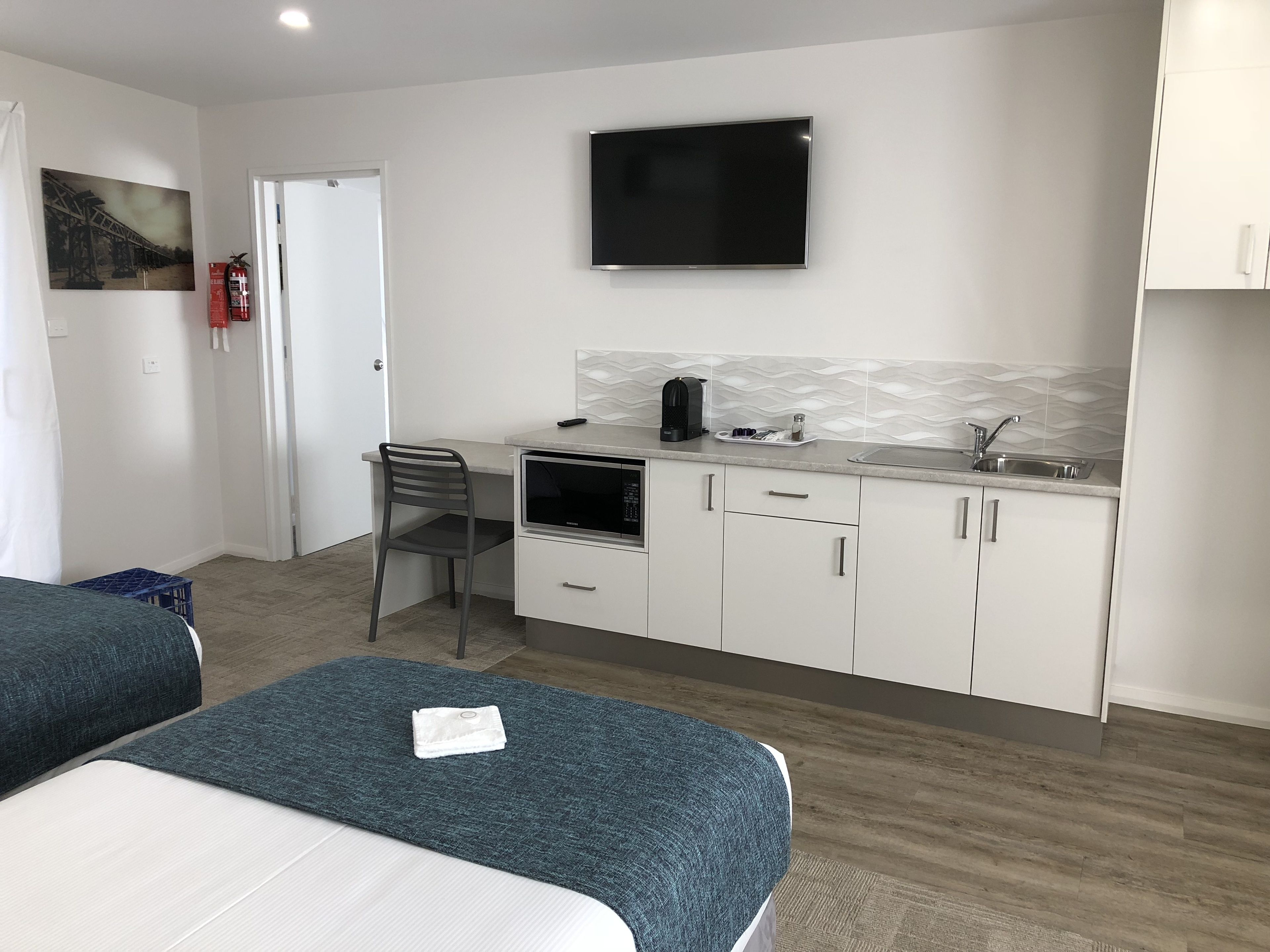 Ein kleines Hotelzimmer mit Küchenzeile, einem Flachbildfernseher, einem Bett mit blauer Bettdecke und einem Sessel.