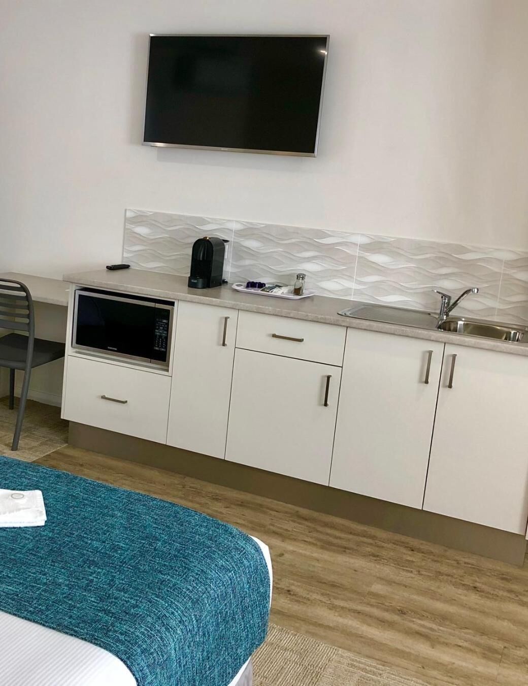 Une chambre d’hôtel compacte dotée d’une kitchenette, d’un téléviseur à écran plat fixé au-dessus du comptoir et d’un lit recouvert d’une couverture bleue.