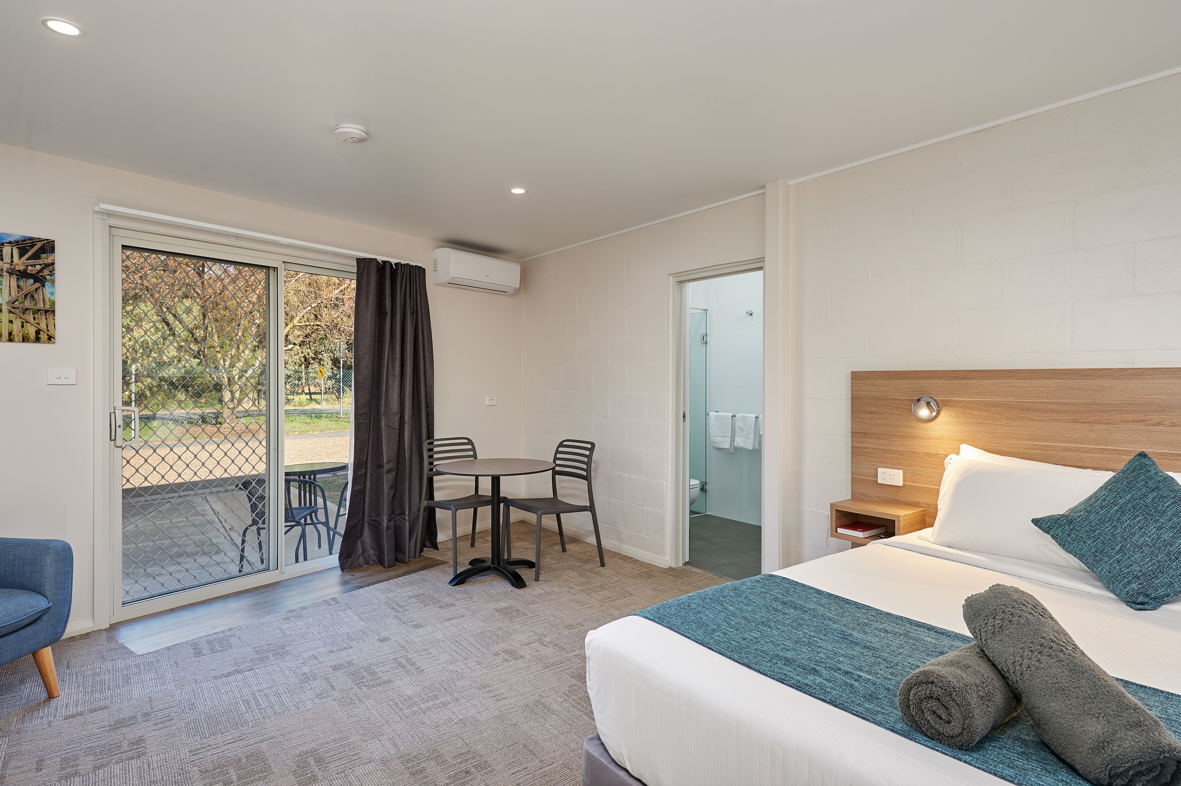 A hotel room with a bed, a table with chairs, a small desk, and a sliding glass door leading to an outdoor area.