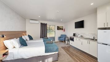 A modern hotel room with a large bed, a desk, a chair, a TV, and a kitchen area.
