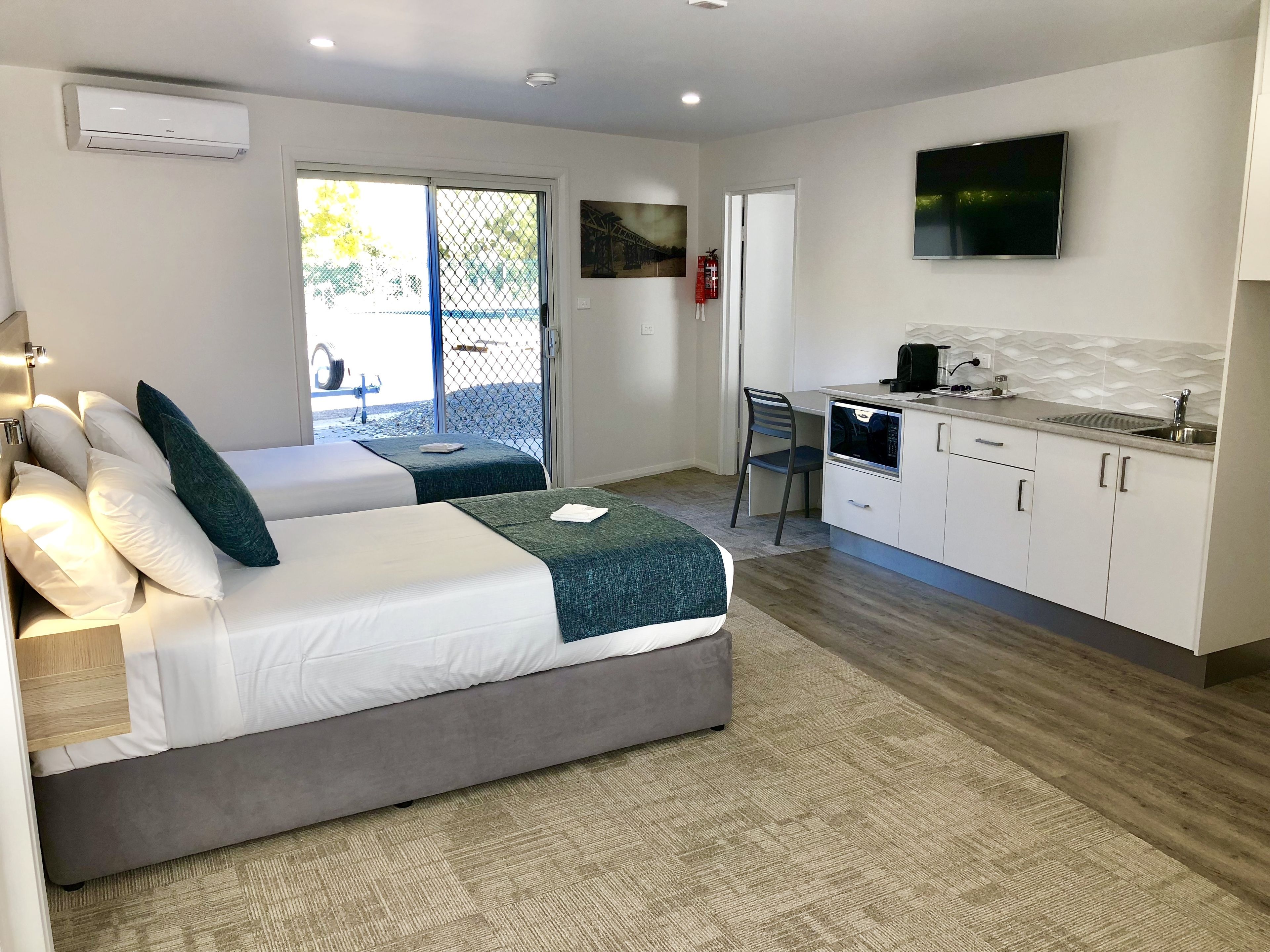 A modern hotel room with a bed, a small kitchenette, and a sliding glass door leading to an outdoor area.