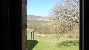 Blick von einem Balkon mit Ziegelwand und Metallgeländer auf eine grüne Landschaft mit einem Baum und sanften Hügeln in der Ferne.