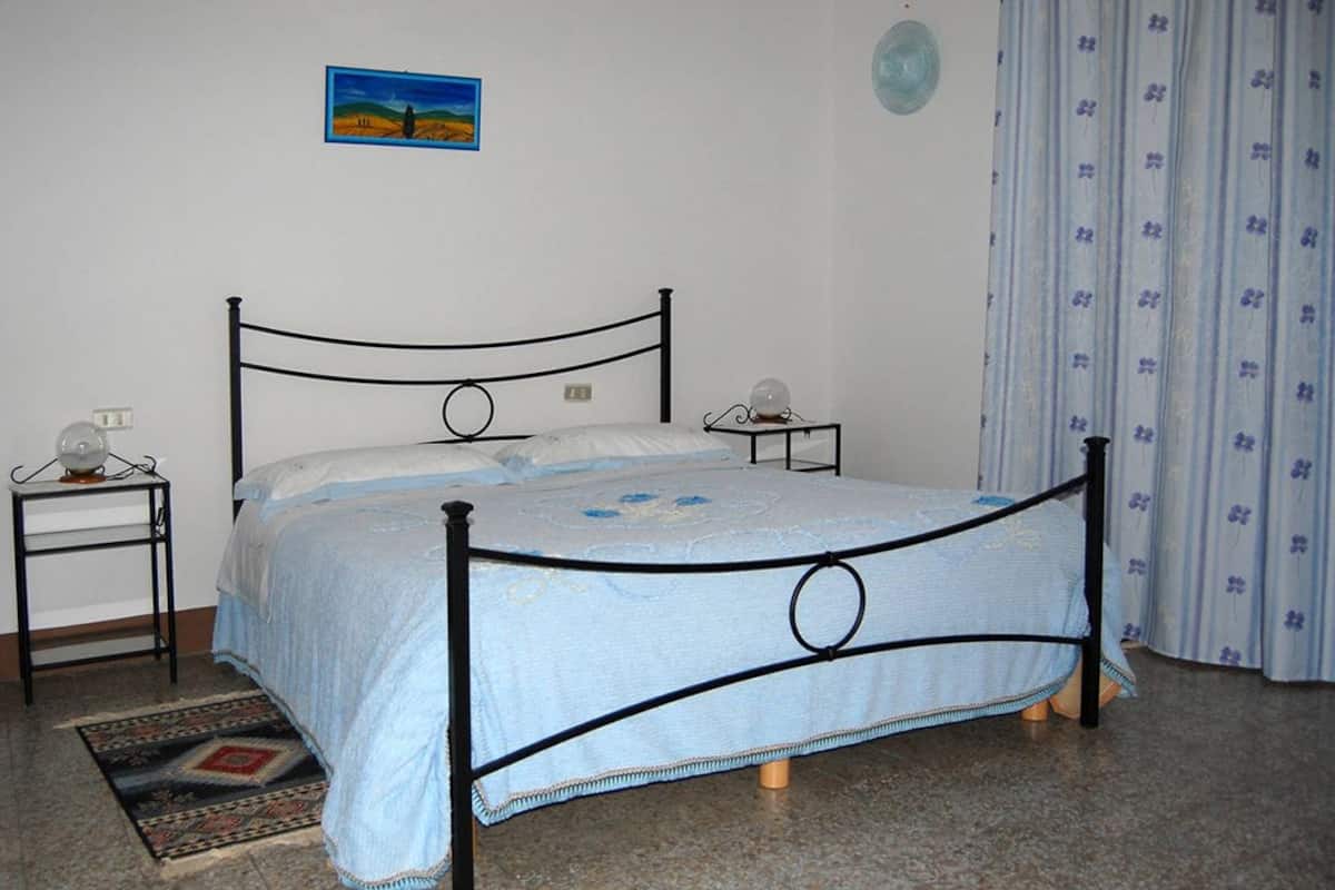 Un dormitorio con una cama, dos mesitas de noche, una lámpara de noche, una alfombra, un cuadro en la pared y una cortina.