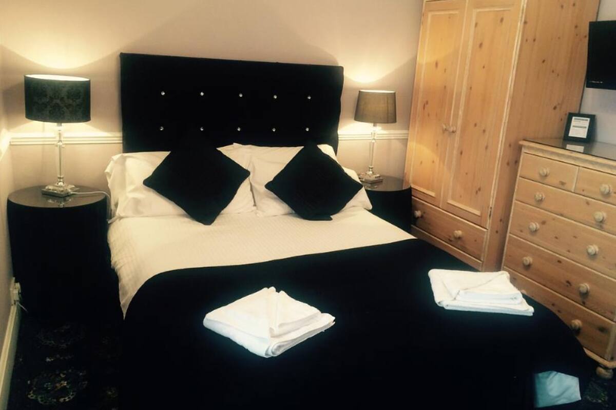 Uma cama bem arrumada com roupa de cama preta e branca, duas luminárias de cabeceira, um aparador de madeira e toalhas brancas dobradas na cama.