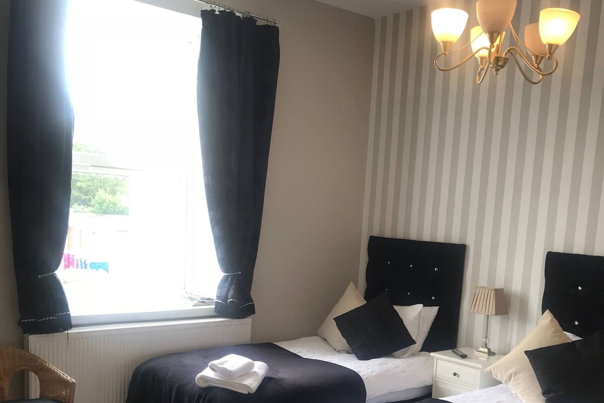 Quarto de hotel com duas camas, janela com cortinas, radiador, cadeira, mesa de cabeceira com abajur e um candelabro.