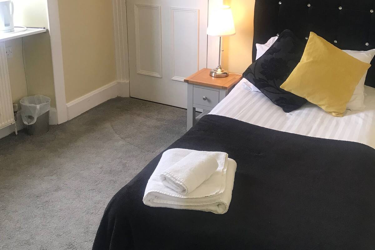 Cama bem arrumada com cabeceira preta, almofadas brancas e amarelas, e uma toalha branca dobrada. Há uma pequena mesa de cabeceira com uma luminária, uma porta branca e um radiador ao fundo.
