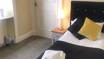 Ein ordentlich bezogenes Bett mit schwarzem Kopfteil, gelb-weißen Kissen und einem gefalteten weißen Handtuch. Ein kleiner Nachttisch mit Lampe, eine weiße Tür und ein Heizkörper sind im Hintergrund zu sehen.