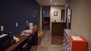 A hotel room with a wooden desk, a microwave, a chair, a red chair, a small table, and a stack of boxes. - POPTONE the Guest House - Hostel (Hiroshima)