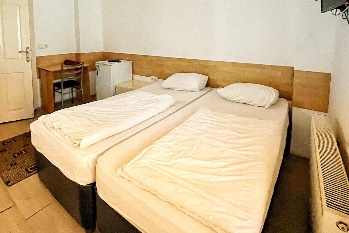 Ein Hotelzimmer mit zwei Einzelbetten, einem Schreibtisch mit Stuhl, einer kleinen Schreibtischlampe und einem an der Wand befestigten Fernseher.