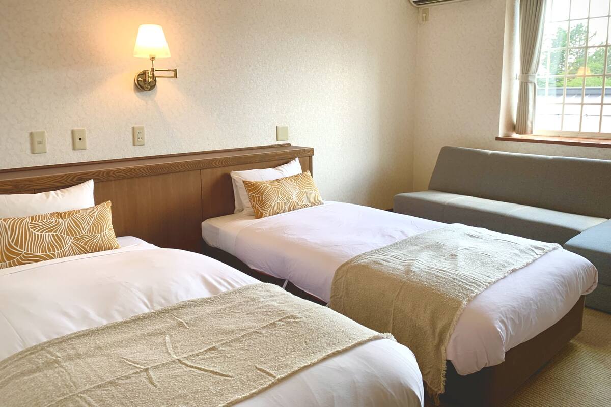 Ein Hotelzimmer mit zwei Einzelbetten, einem Schlafsofa, einer Wandlampe und einem Fenster mit Vorhängen.